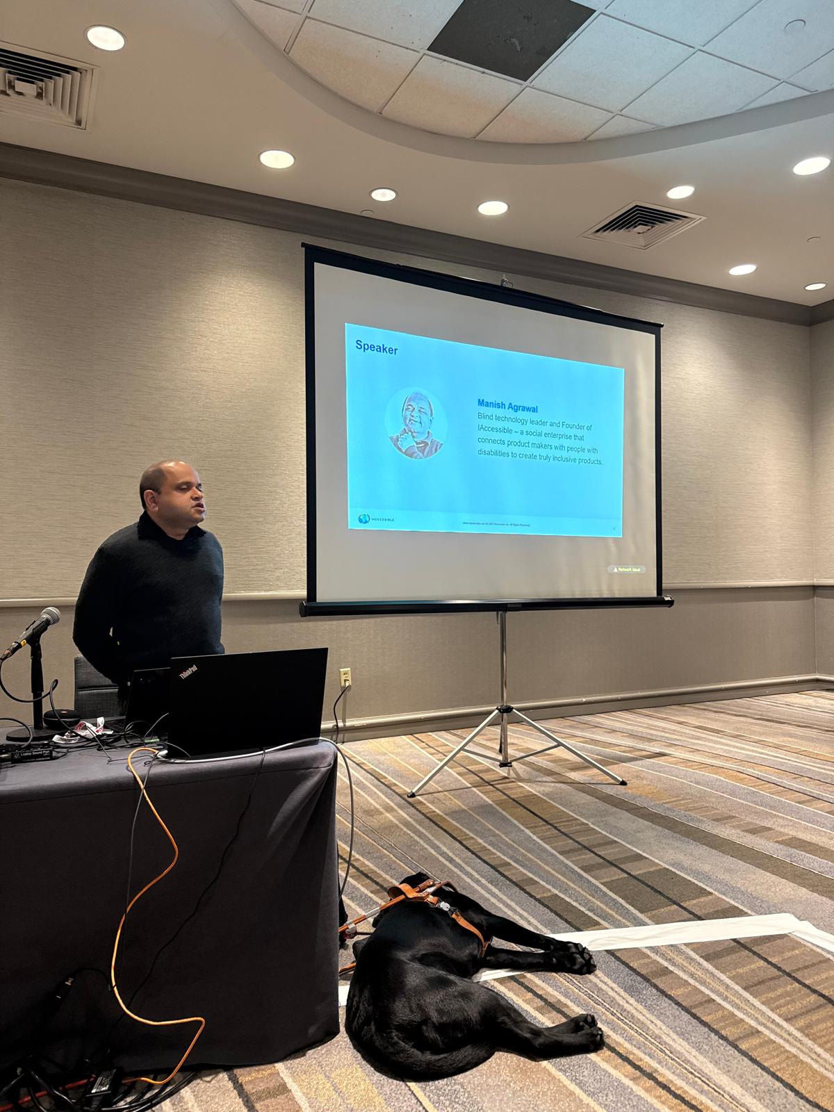 Manish standing in front of the presentation with the black labrador Lenny sleeping on the floor with his back to the camera